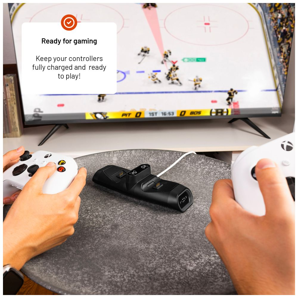 Mani che tengono i controller vicino a una stazione di ricarica, con una TV che mostra una partita di hockey.