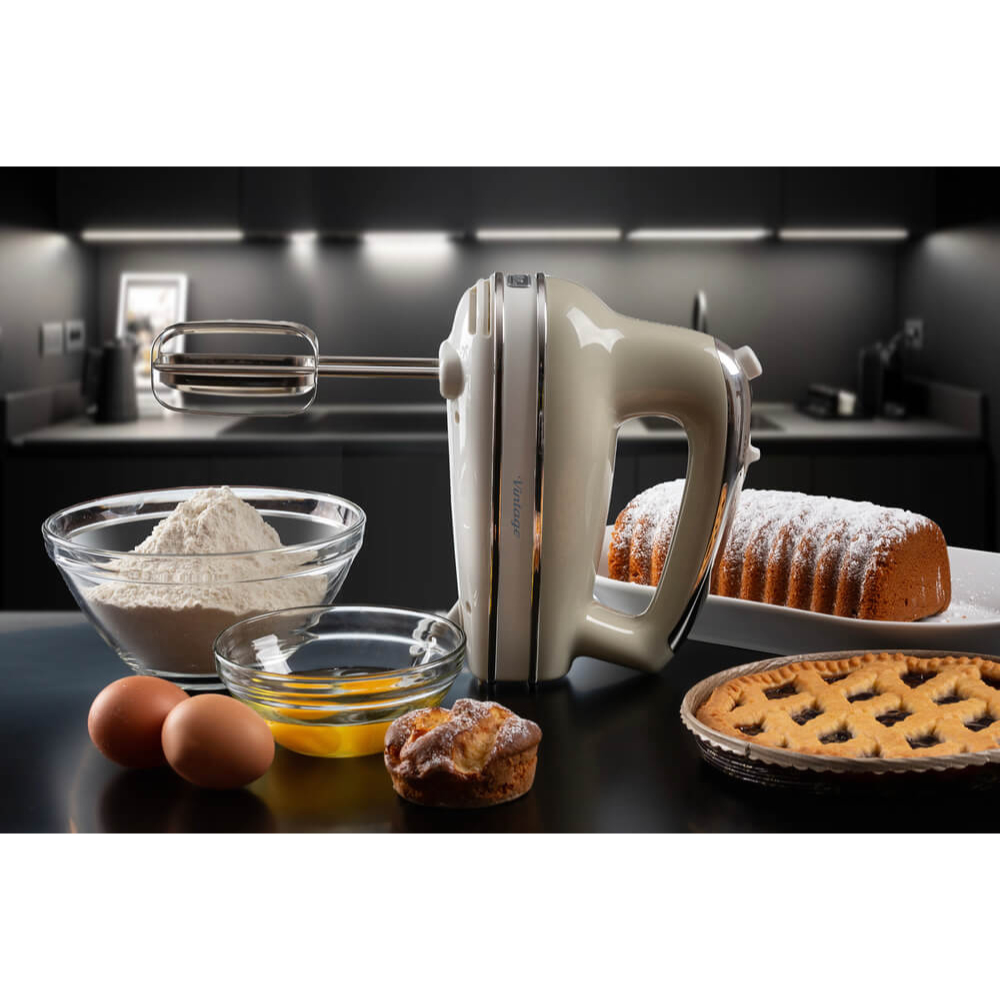 Küchenszene mit beigefarbenem Handmixer, Schüsseln mit Zutaten und Backwaren auf dunkler Oberfläche. Hintergrund ist eine Küche.