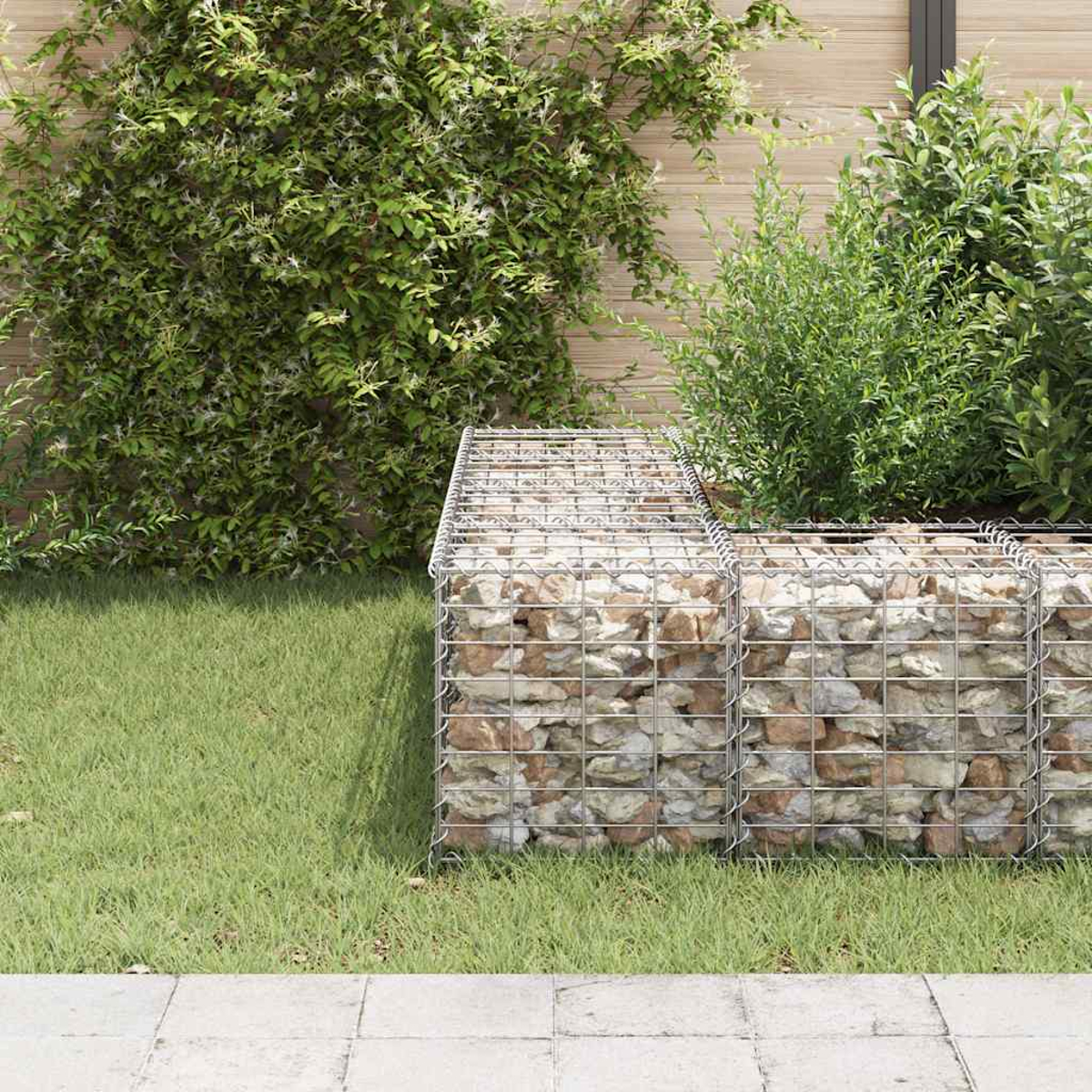 Eine Gartenszene mit einer Gabionenmauer, die mit Steinen gefüllt ist. Grünes Gras und Pflanzen umgeben die Mauer. Ein Steinweg davor.