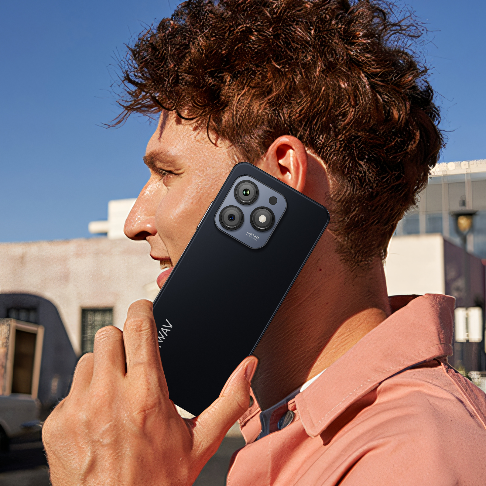 Mężczyzna trzymający czarny telefon z potrójnym aparatem przy uchu. Tło niebieskiego nieba. Ma na sobie różową koszulę.