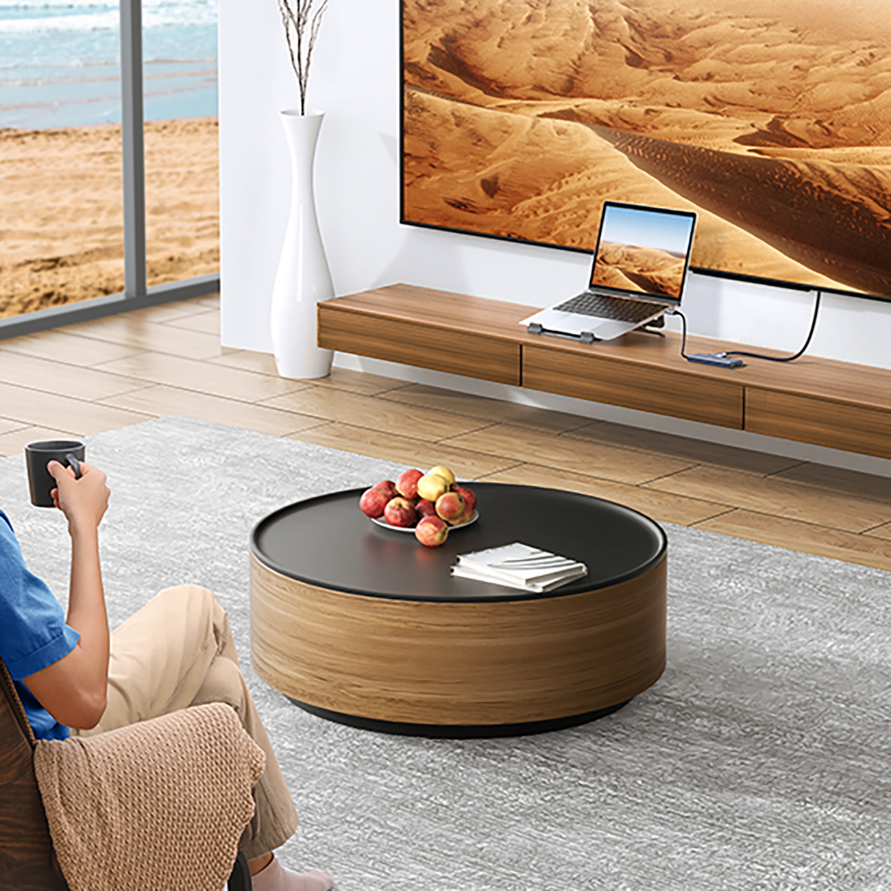 Osoba siedząca na krześle trzymająca kubek, w pobliżu okrągłego stołu z owocami. Laptop jest na stojaku telewizyjnym.