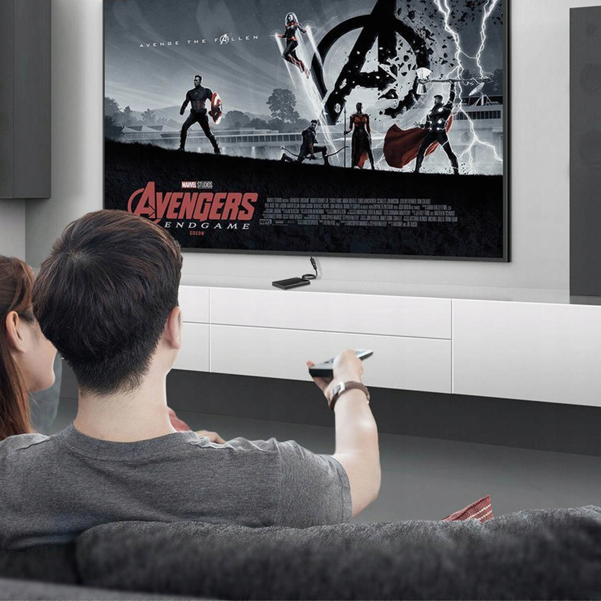 Dwie osoby oglądają film w telewizji. Telewizor jest zamontowany na białej szafce. Film to Avengers.