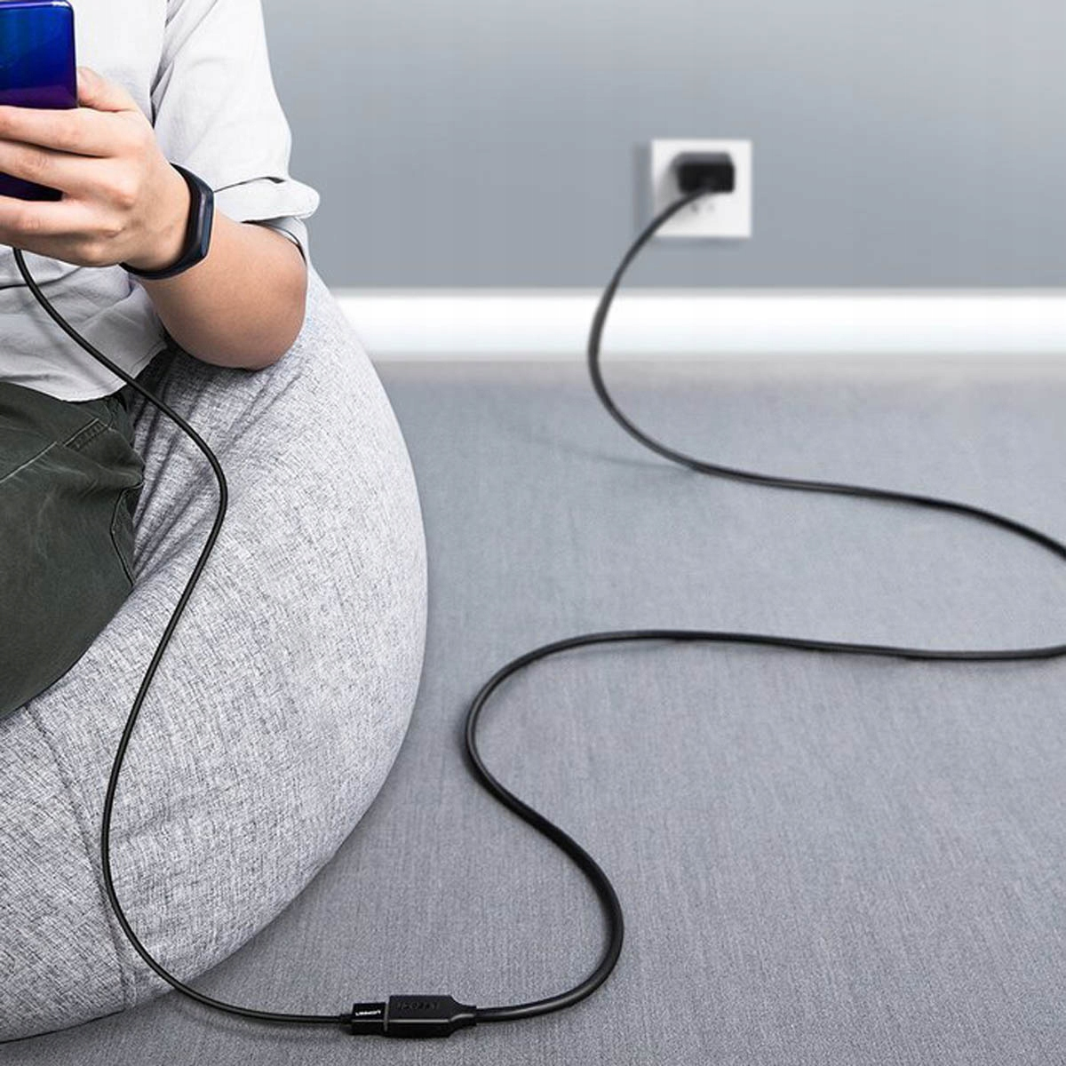 Osoba siedząca na worku sako ładuje telefon kablem UGREEN podłączonym do gniazdka ściennego.