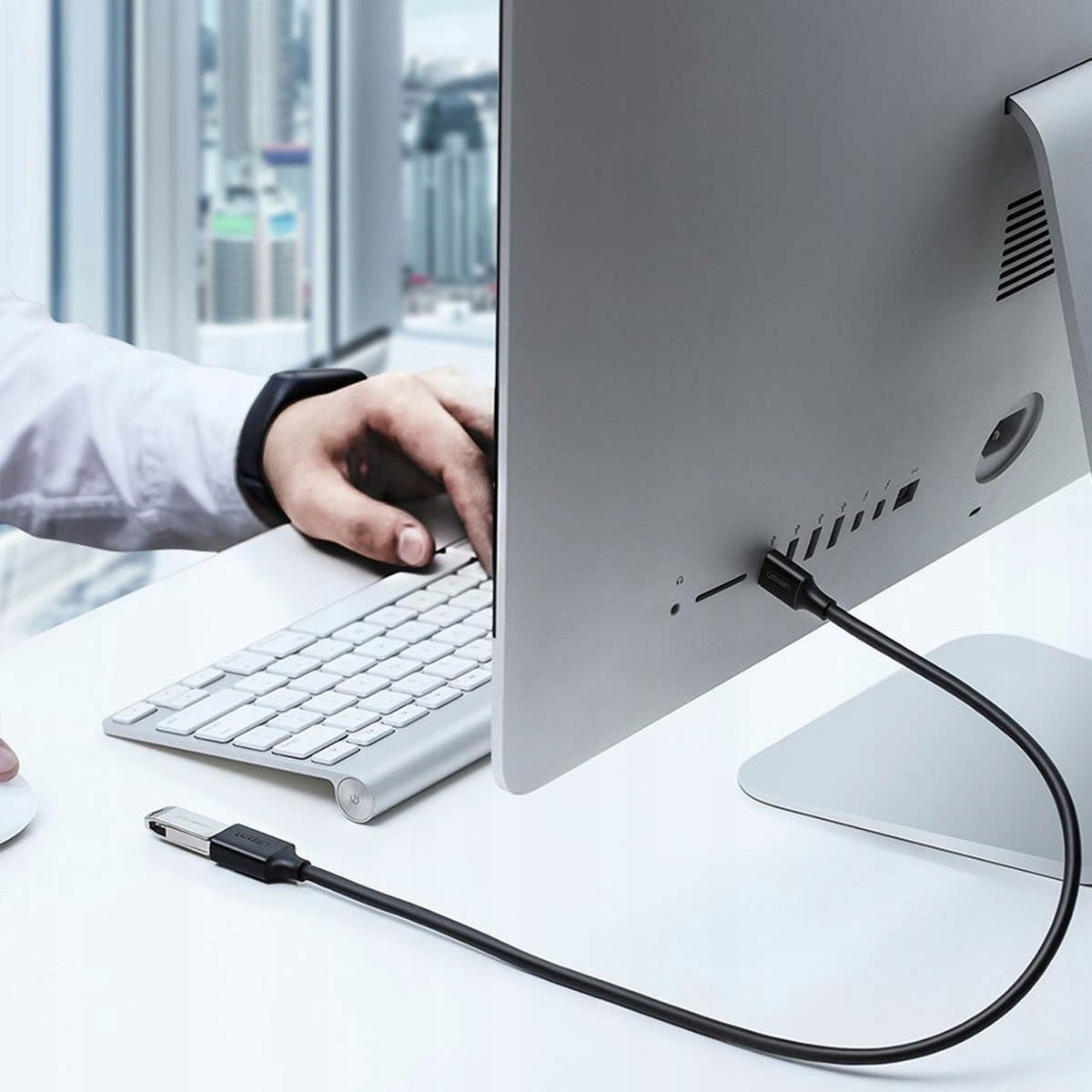 Osoba korzystająca z laptopa i monitora z czarnym kablem USB podłączonym z tyłu, na białym biurku.