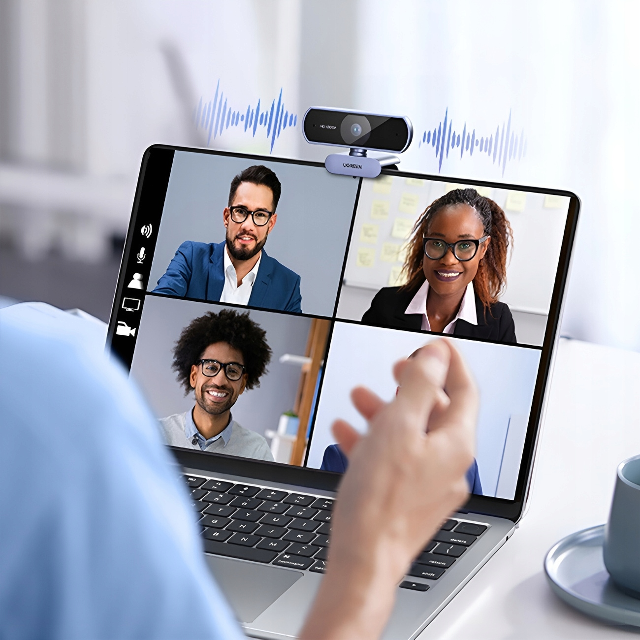 Laptop z kamerą internetową na górze, pokazujący połączenie wideo z czterema osobami. Ręka dotyka laptopa.