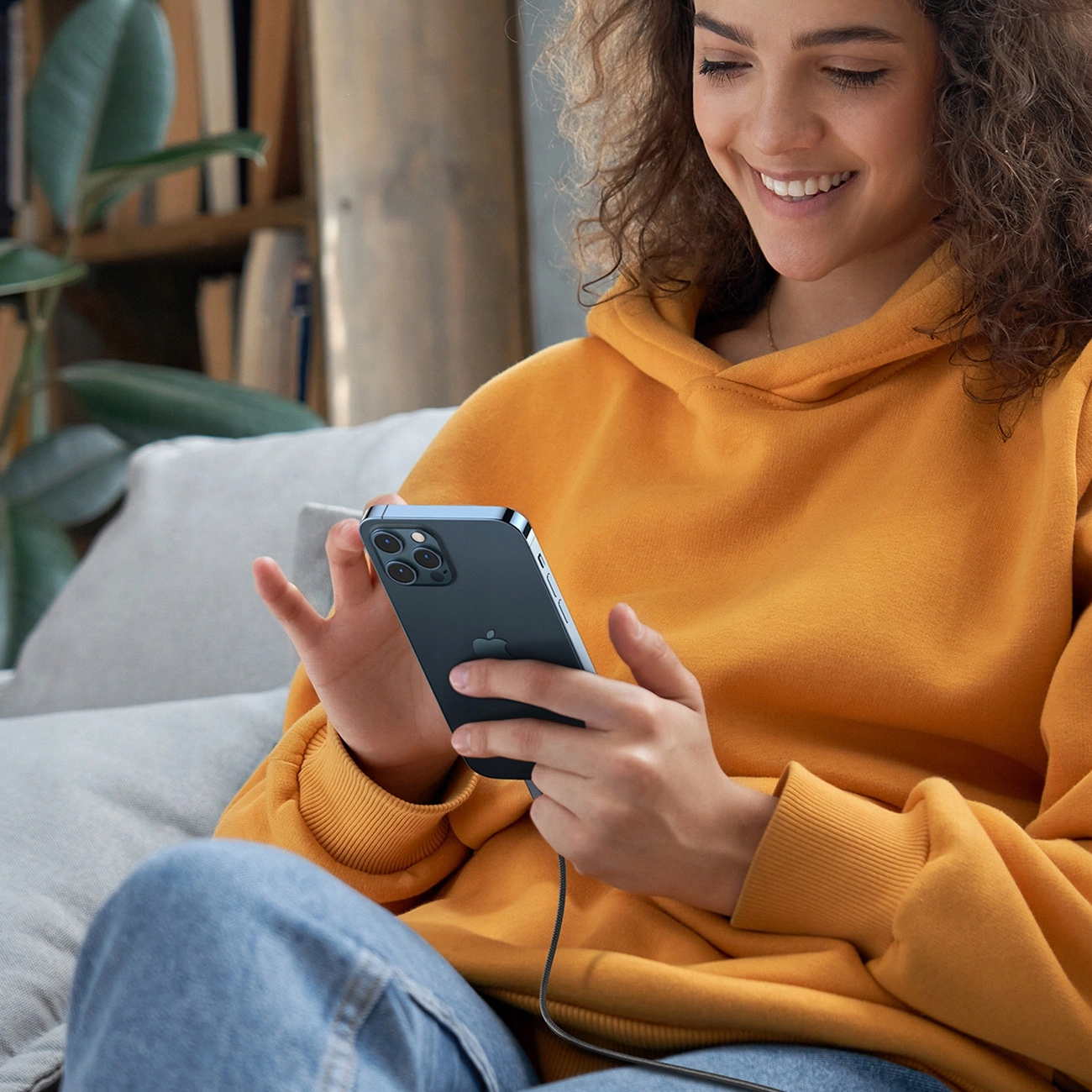 Kobieta w pomarańczowej bluzie z kapturem trzyma telefon, uśmiecha się. Siedzi na kanapie. W tle półka na książki.