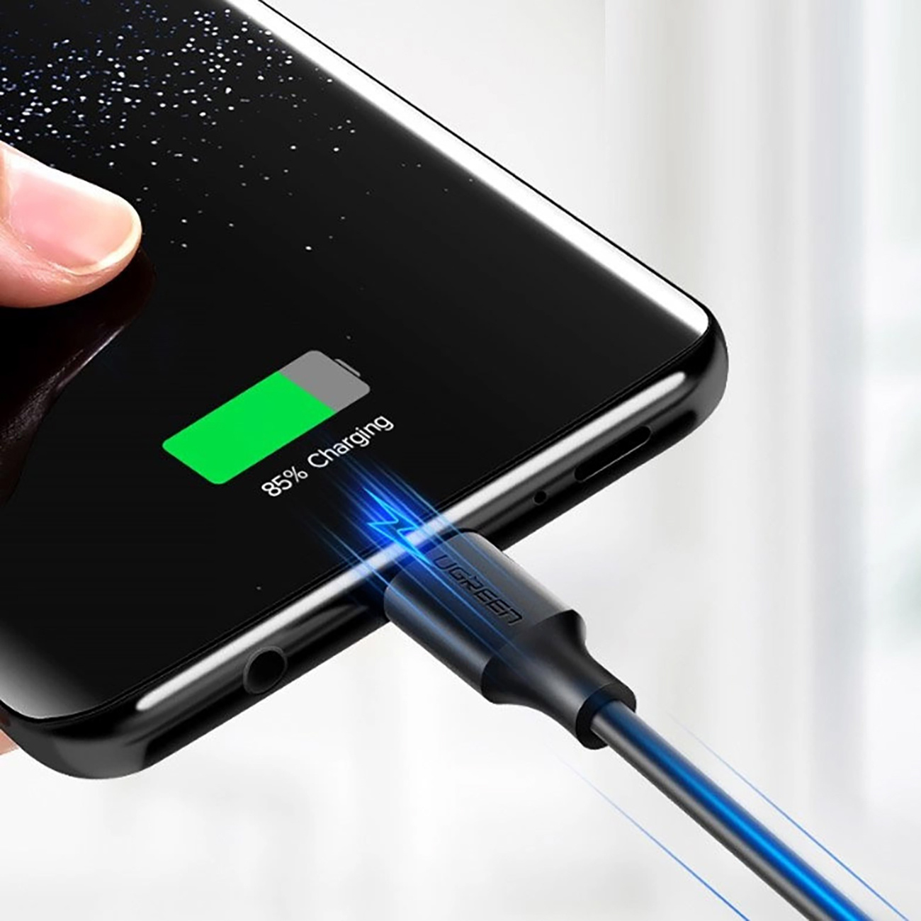 Czarny telefon ładuje się kablem UGREEN. Ekran pokazuje 85% naładowania. Ręka trzyma telefon.