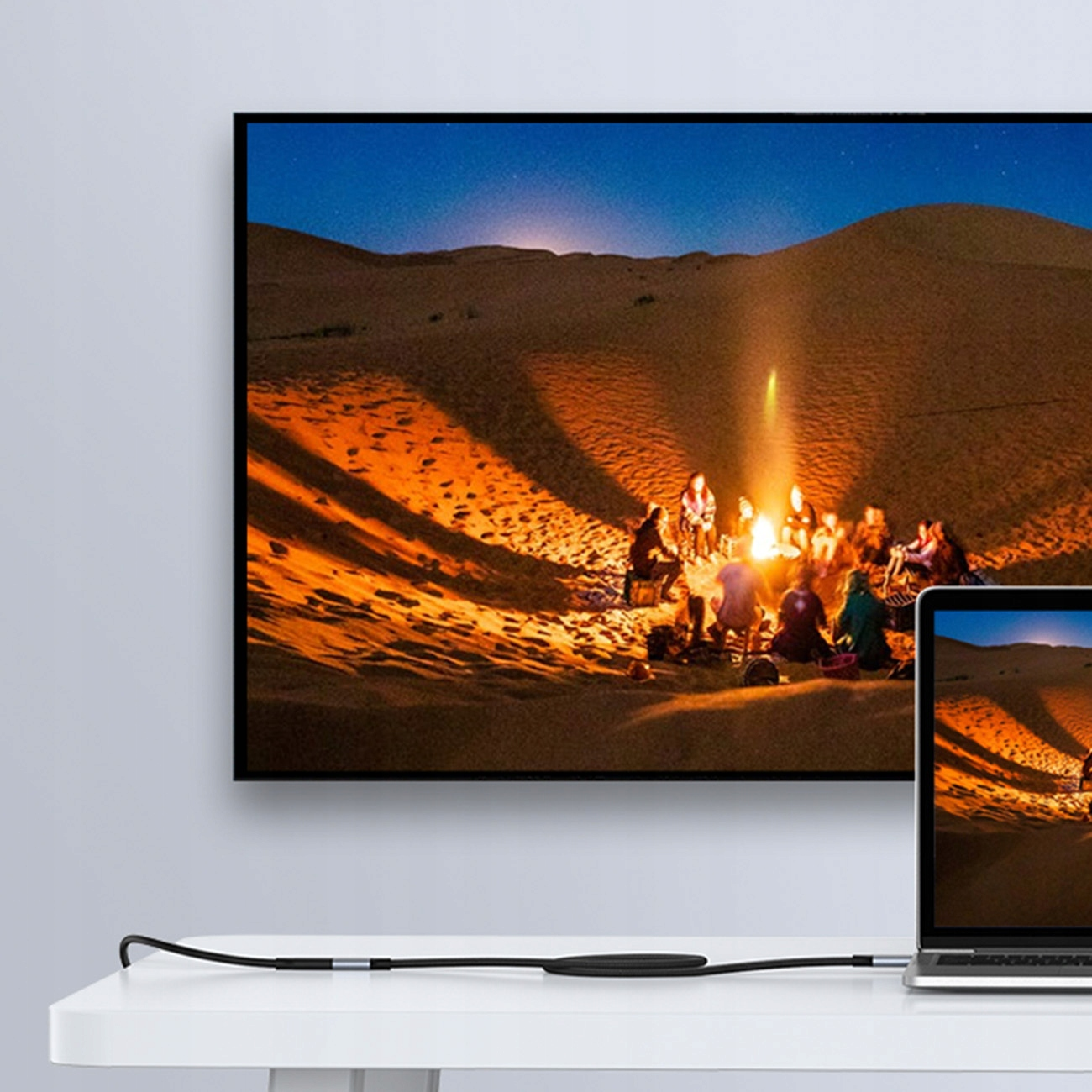 Una TV mostra una scena del deserto con un falò. Un laptop e un cavo di ricarica sono su un tavolo.