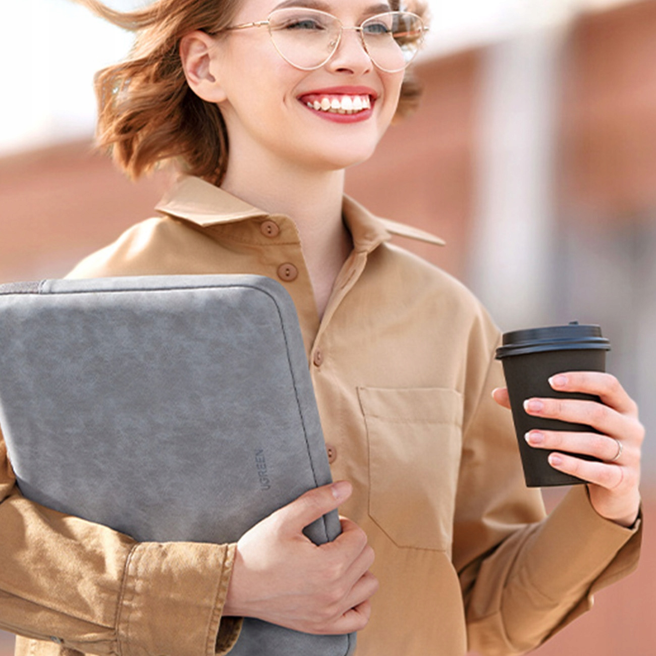 Kobieta trzymająca szarą torbę na laptopa i filiżankę kawy. Uśmiecha się, z rozmytym tłem.