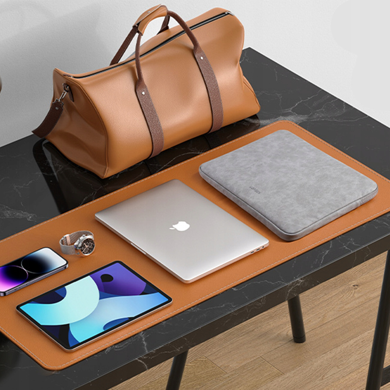 Konfiguracja biurka z różnymi elementami: laptopem, tabletem, telefonem, zegarkiem, torbą i szarą torbą na laptopa na skórzanej macie.