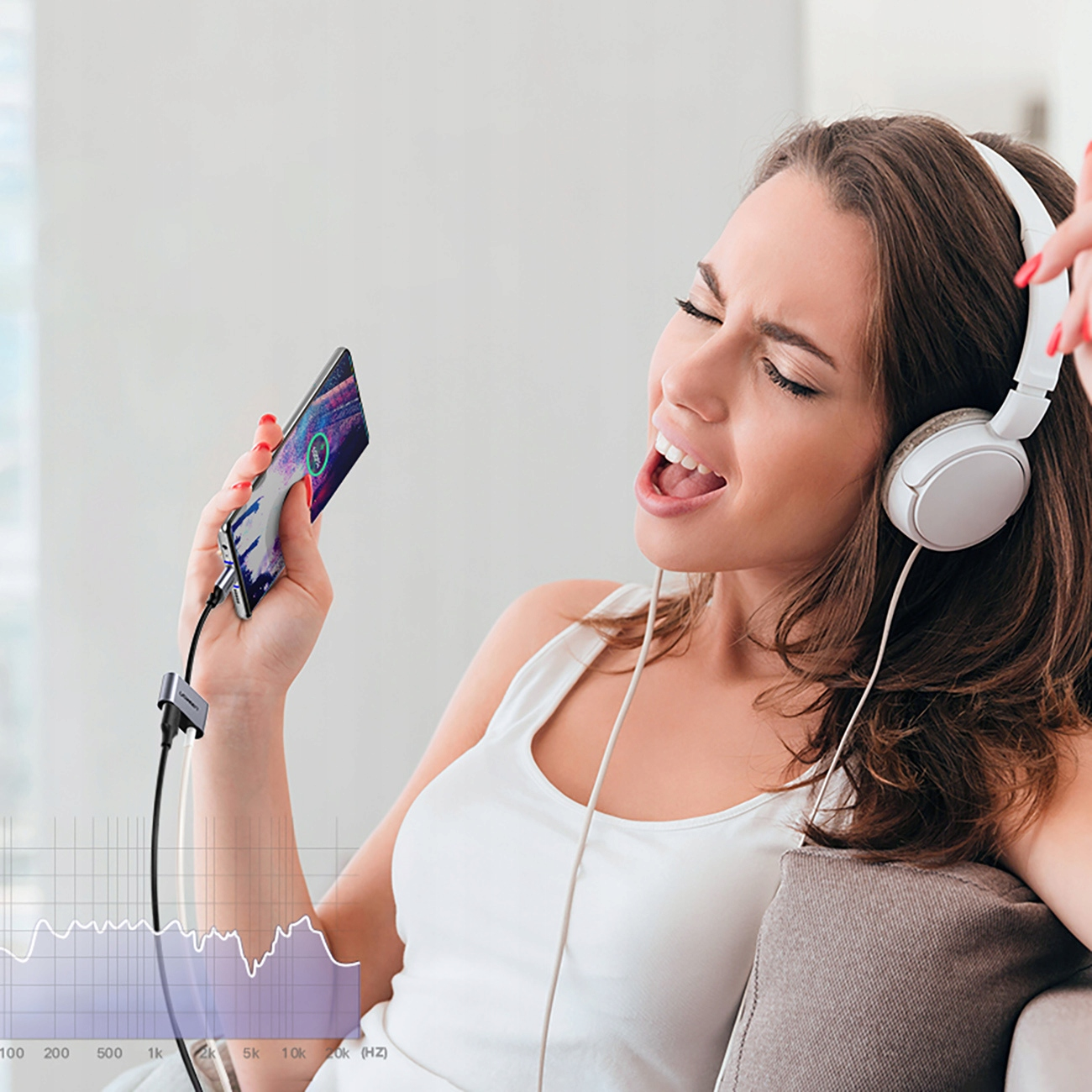 Kobieta śpiewa ze słuchawkami na uszach, trzymając telefon podłączony do kabla adaptera słuchawkowego, z wykresem audio.