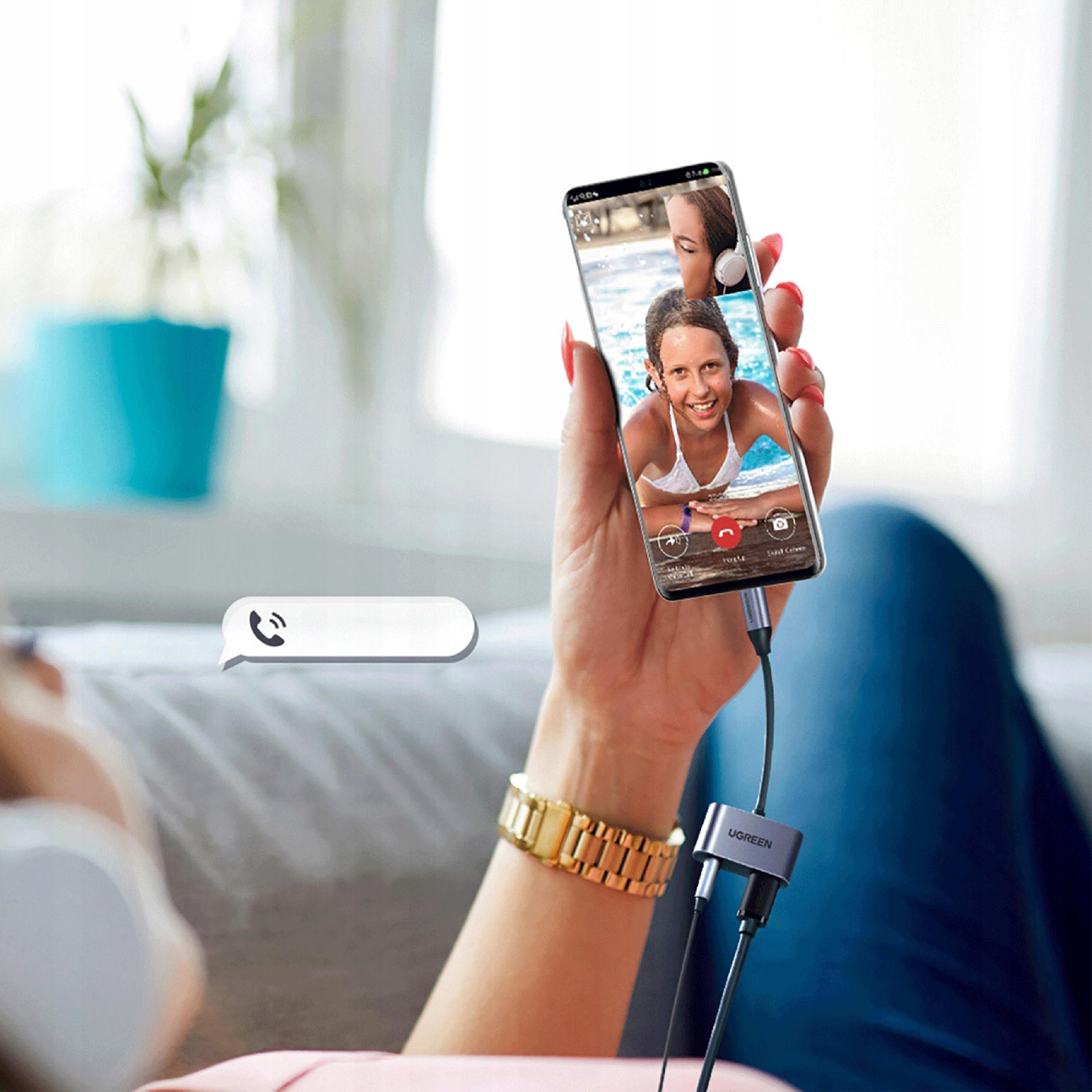 Osoba prowadzi rozmowę wideo na telefonie. Srebrny adapter łączy telefon ze słuchawkami. Kobieta siedzi na kanapie.