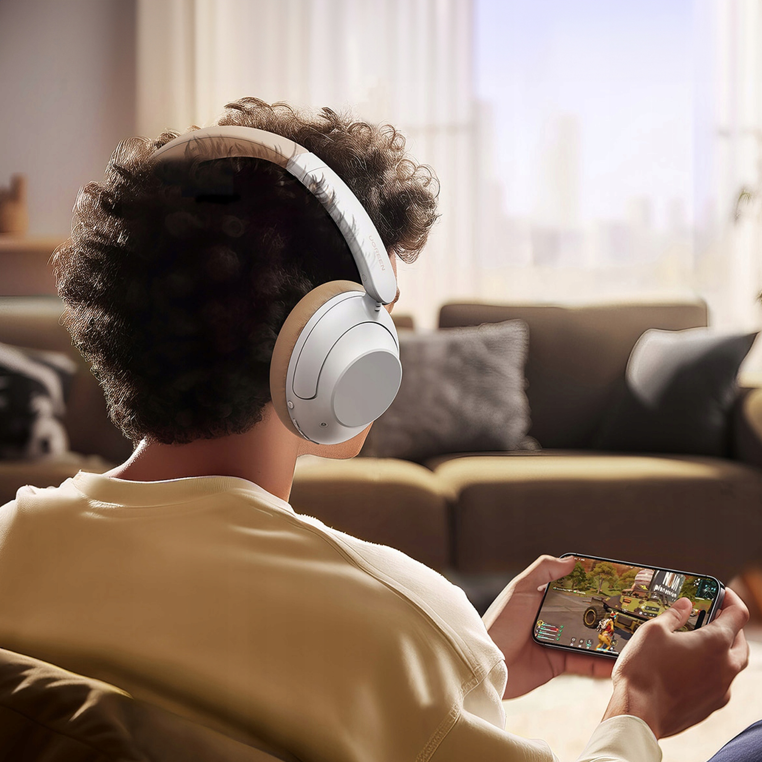 Osoba w słuchawkach gra w grę na telefonie. Brązowa sofa w tle.