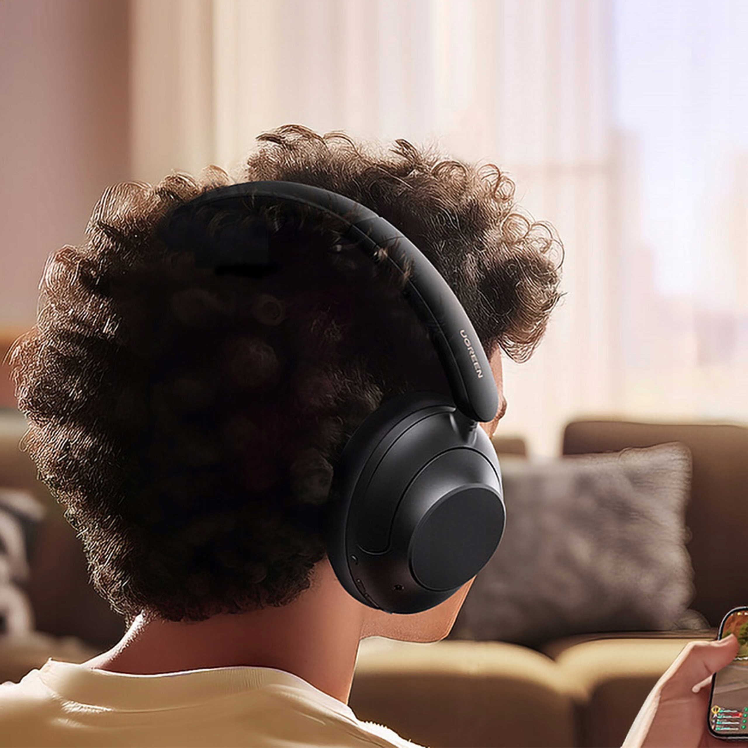 Osoba w czarnych słuchawkach UGREEN patrzy na telefon pokazujący grę, brązowa sofa w tle.