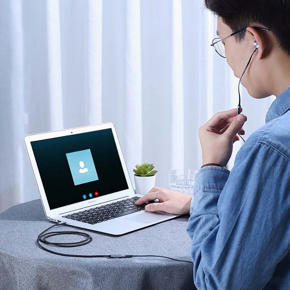 Mężczyzna w okularach używa laptopa i słuchawek. Mała roślina na stole. Okno z zasłoną w tle.