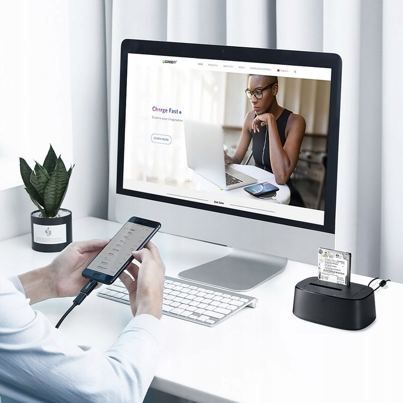 Eine Person benutzt ein Telefon und einen Computer. Eine Festplatten-Dockingstation steht auf dem Tisch, der Bildschirm zeigt eine Website.