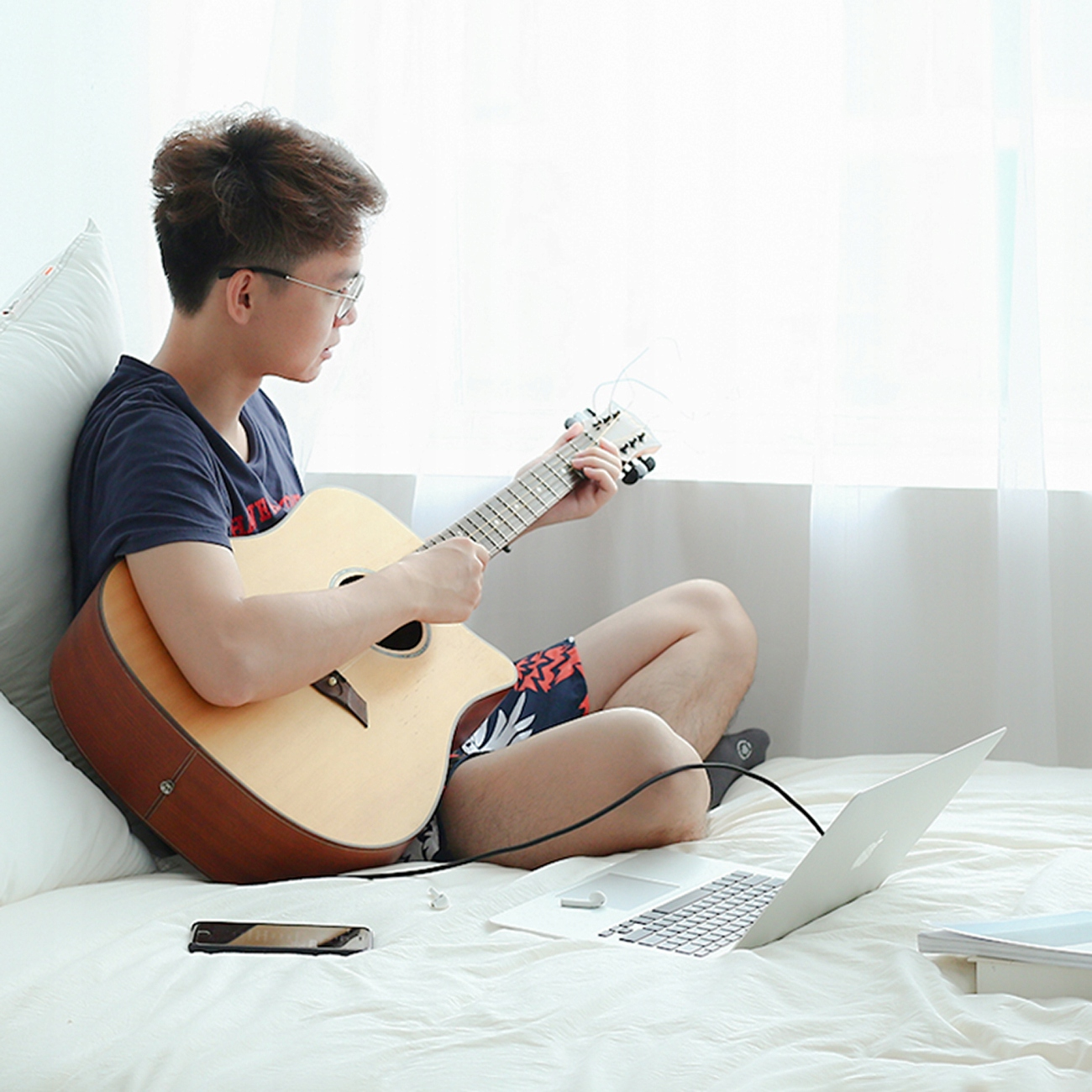 Mężczyzna siedzi na łóżku, gra na gitarze i używa laptopa ze słuchawkami. Tło to okno.