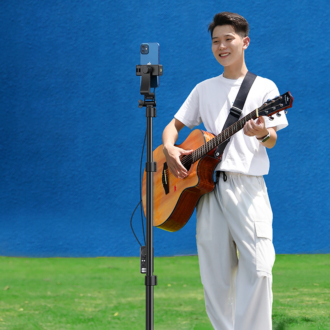 Mężczyzna gra na gitarze, z telefonem zamontowanym na statywie. Tło jest niebieskie.
