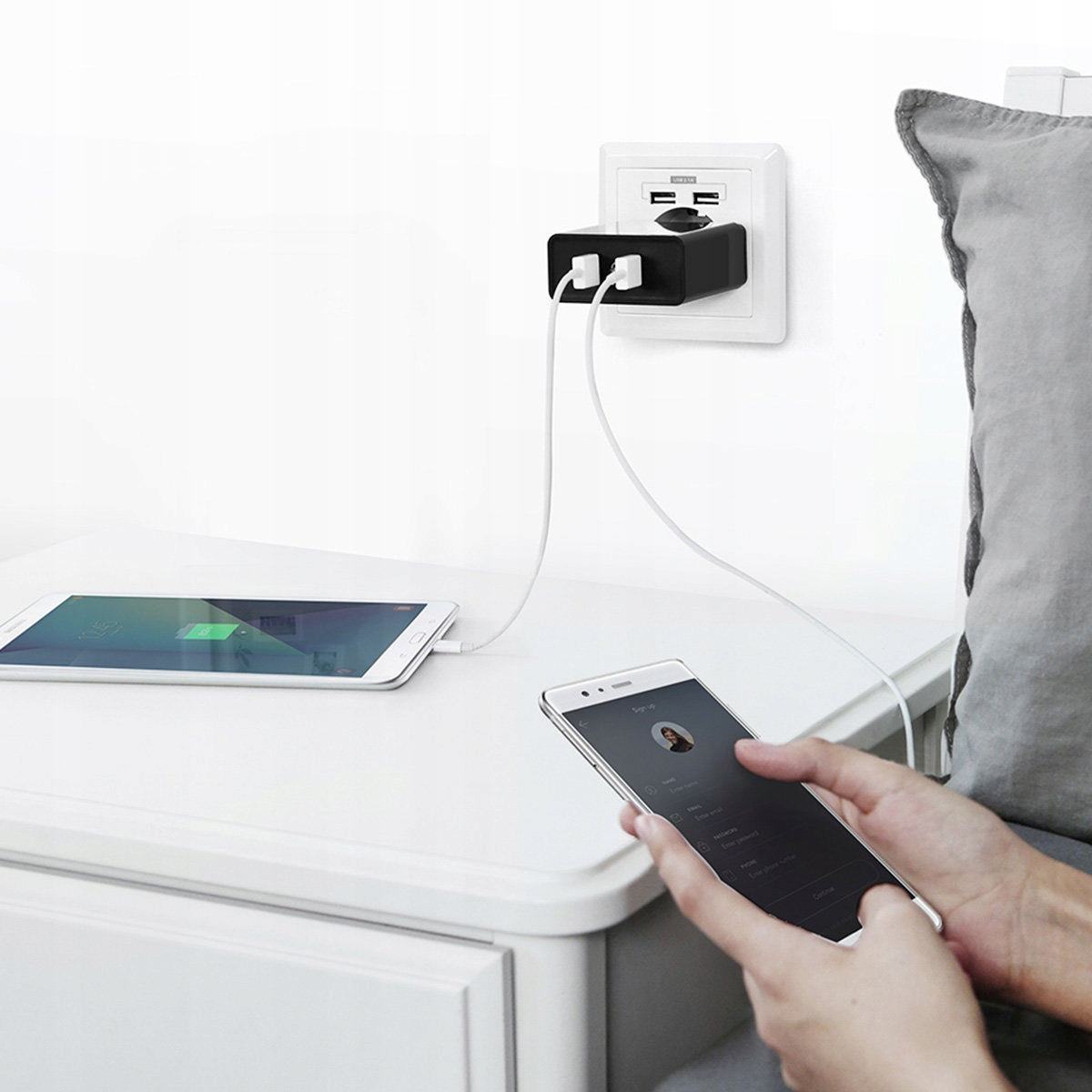 Ein Tablet und ein Telefon werden über ein schwarzes Netzteil an einer weißen Wand aufgeladen.