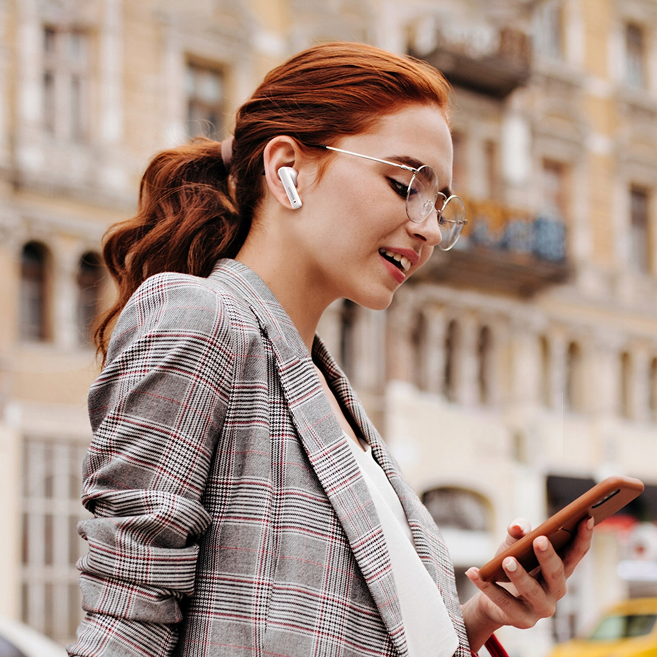 Kobieta z rudymi włosami w okularach i słuchawkami, trzymająca telefon, na zewnątrz w kurtce w kratę.