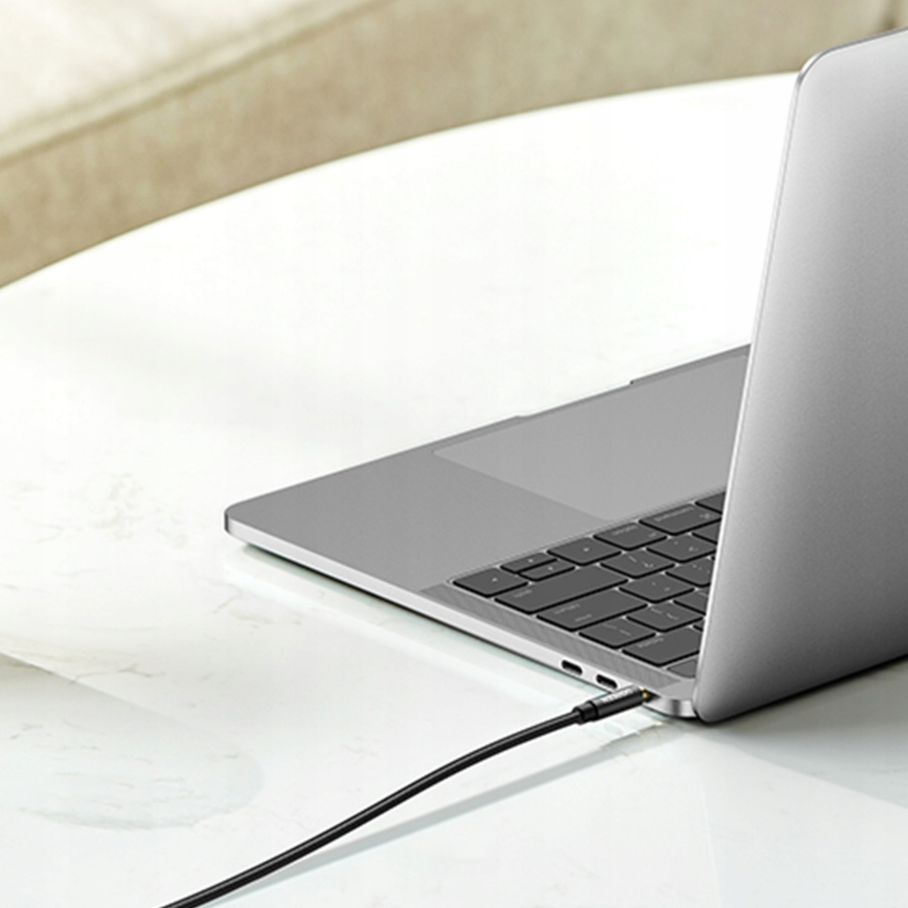 Laptop z podłączonym kablem do ładowania. Kabel jest czarny, a laptop stoi na białym stole. Proste otoczenie.