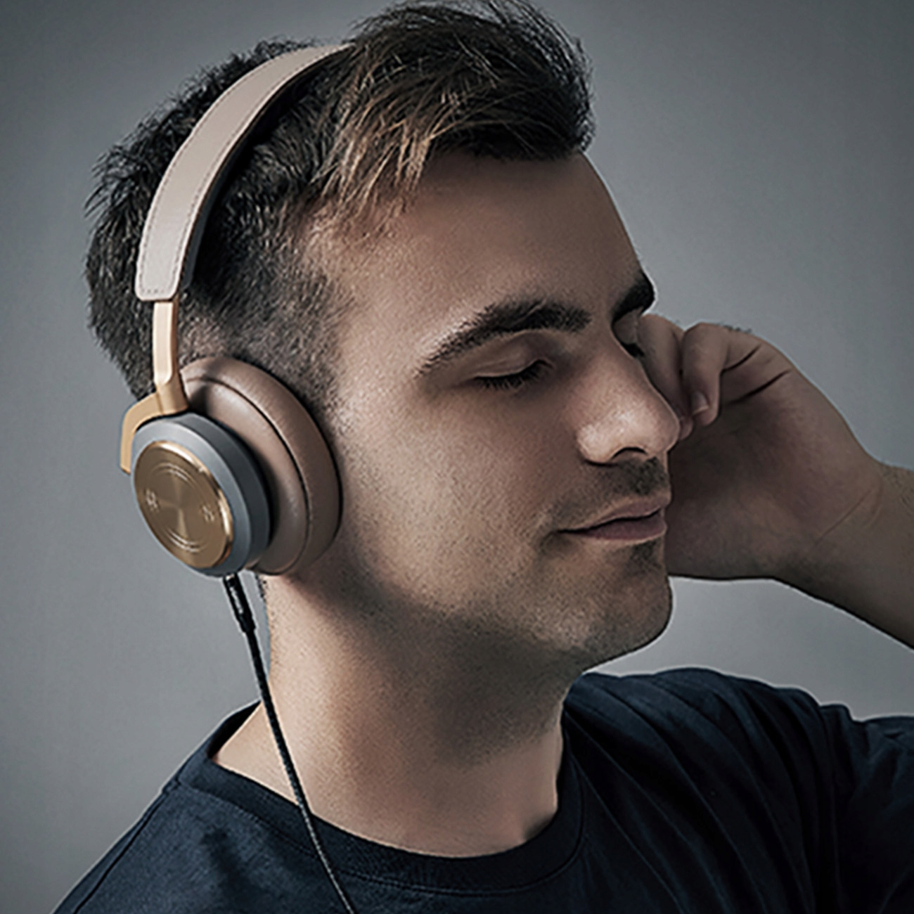 Mężczyzna w brązowo-szarych słuchawkach, z zamkniętymi oczami, dotykający ucha dłonią. Ma na sobie granatową koszulę.