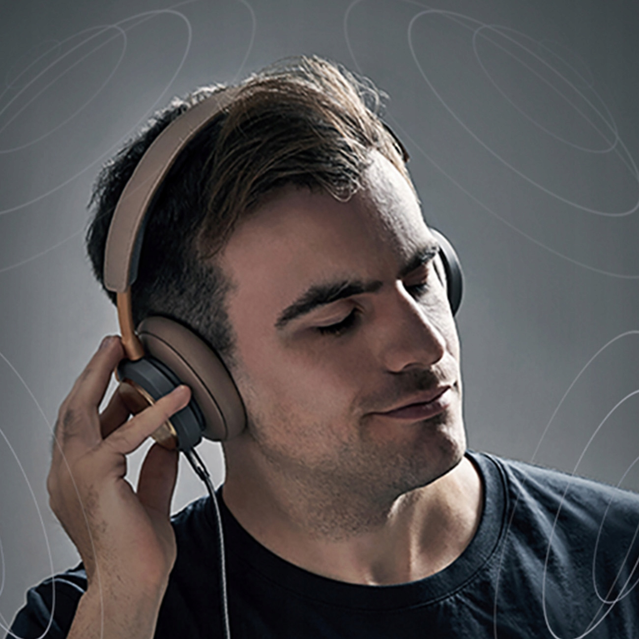 Mężczyzna w słuchawkach, patrzący w dół, uśmiechnięty. Szare tło z jasnymi okręgami.