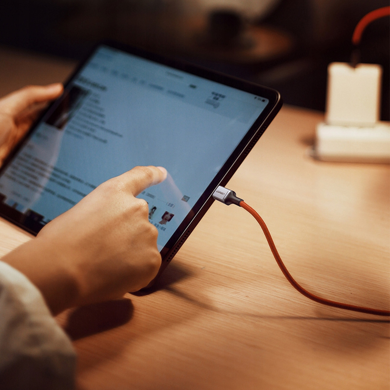 Osoba korzysta z tabletu, podłączonego czerwonym kablem. Tablet leży na drewnianej powierzchni.