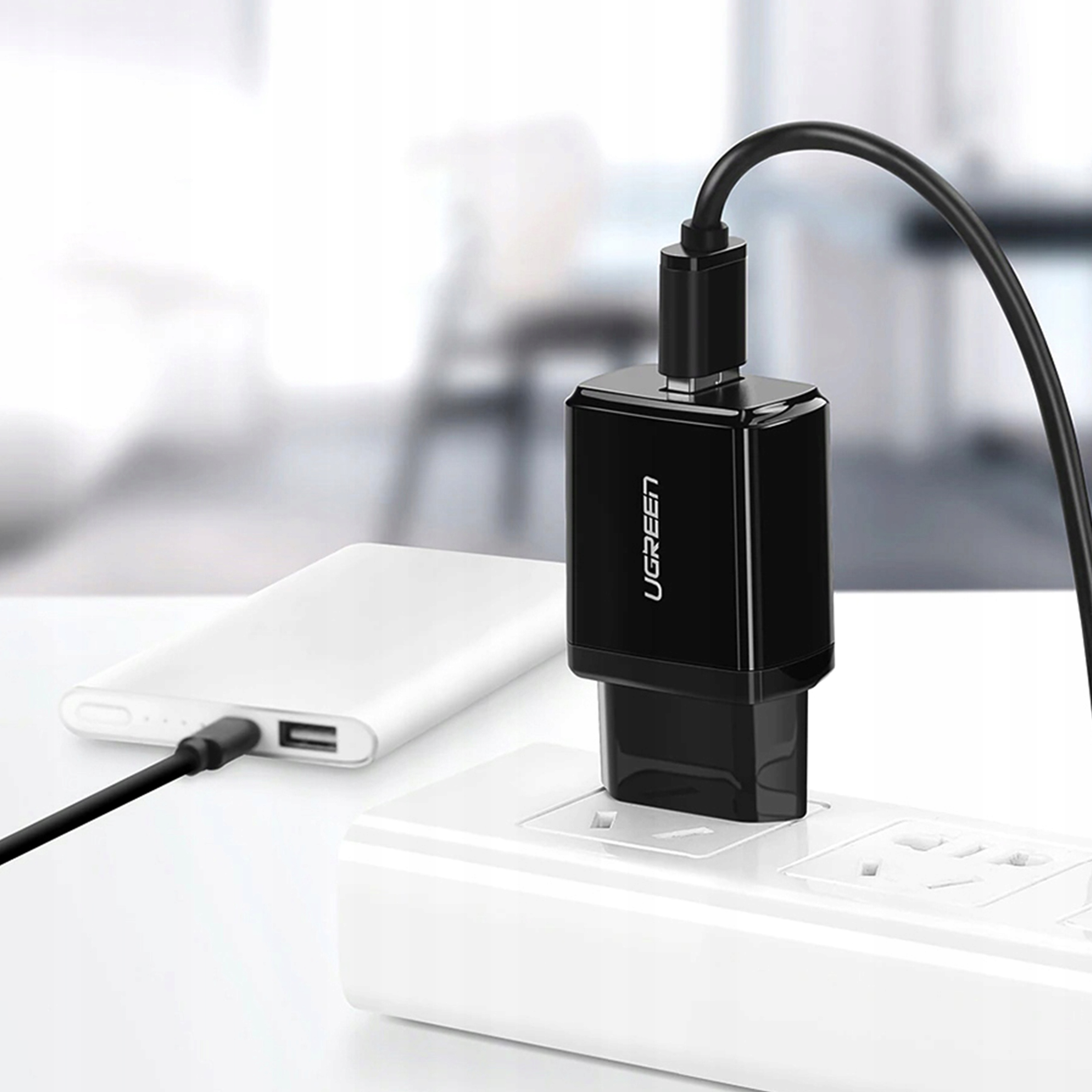 Czarna ładowarka UGREEN podłączona do gniazdka ściennego, ładująca power bank. Biała powierzchnia znajduje się na pierwszym planie.