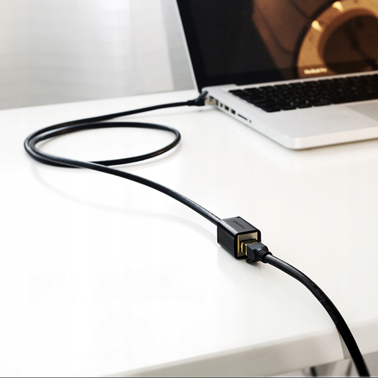 Laptop z podłączonym czarnym kablem USB. Kabel leży na białym stole.