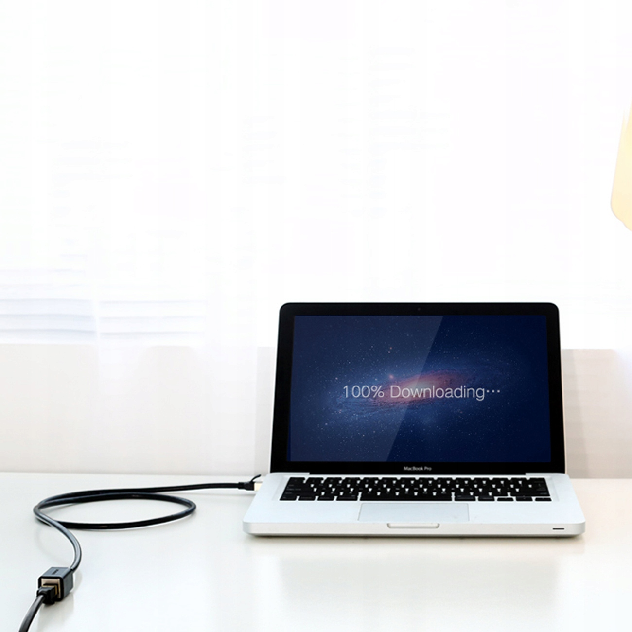 Ekran laptopa wyświetla 100% pobierania. Czarny kabel jest podłączony. Tło jest rozmyte.
