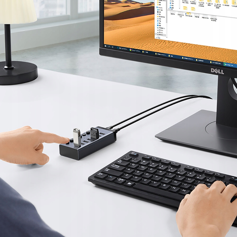 Ręka naciska koncentrator USB na białym biurku. Czarnej klawiatury i monitor komputerowy są w tle.