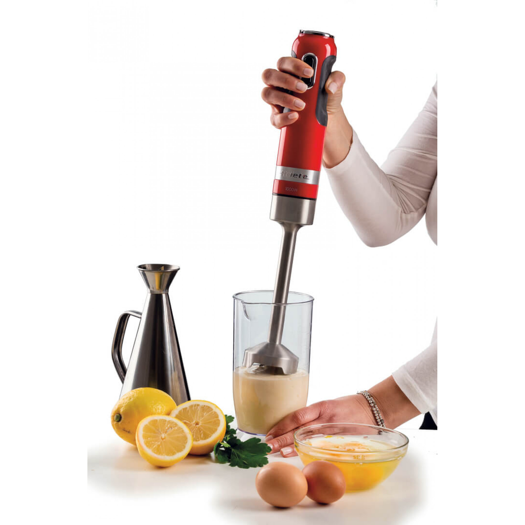 Ein roter Mixer vermischt eine weiße Sauce in einem durchsichtigen Becher. Eier, Zitronen, Petersilie und eine Ölkanne stehen auf dem Tisch.