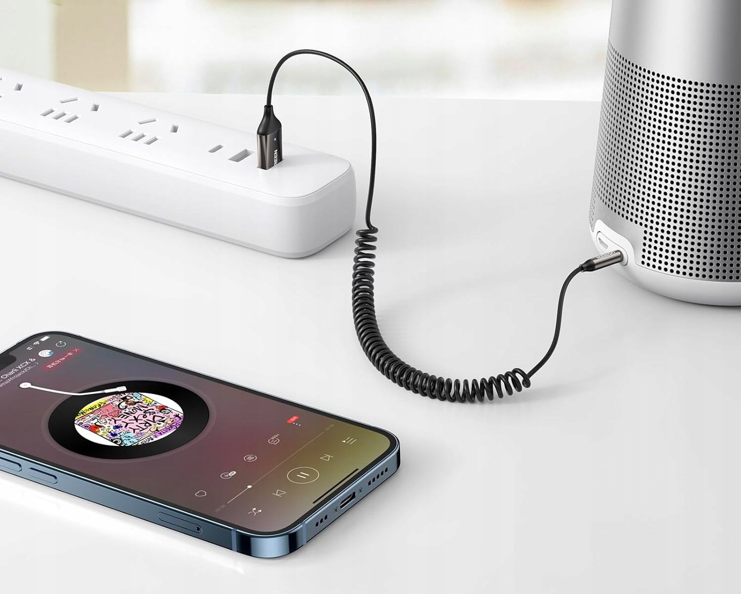 Telefon z aplikacją muzyczną jest podłączony do głośnika i listwy zasilającej za pomocą czarnych kabli.