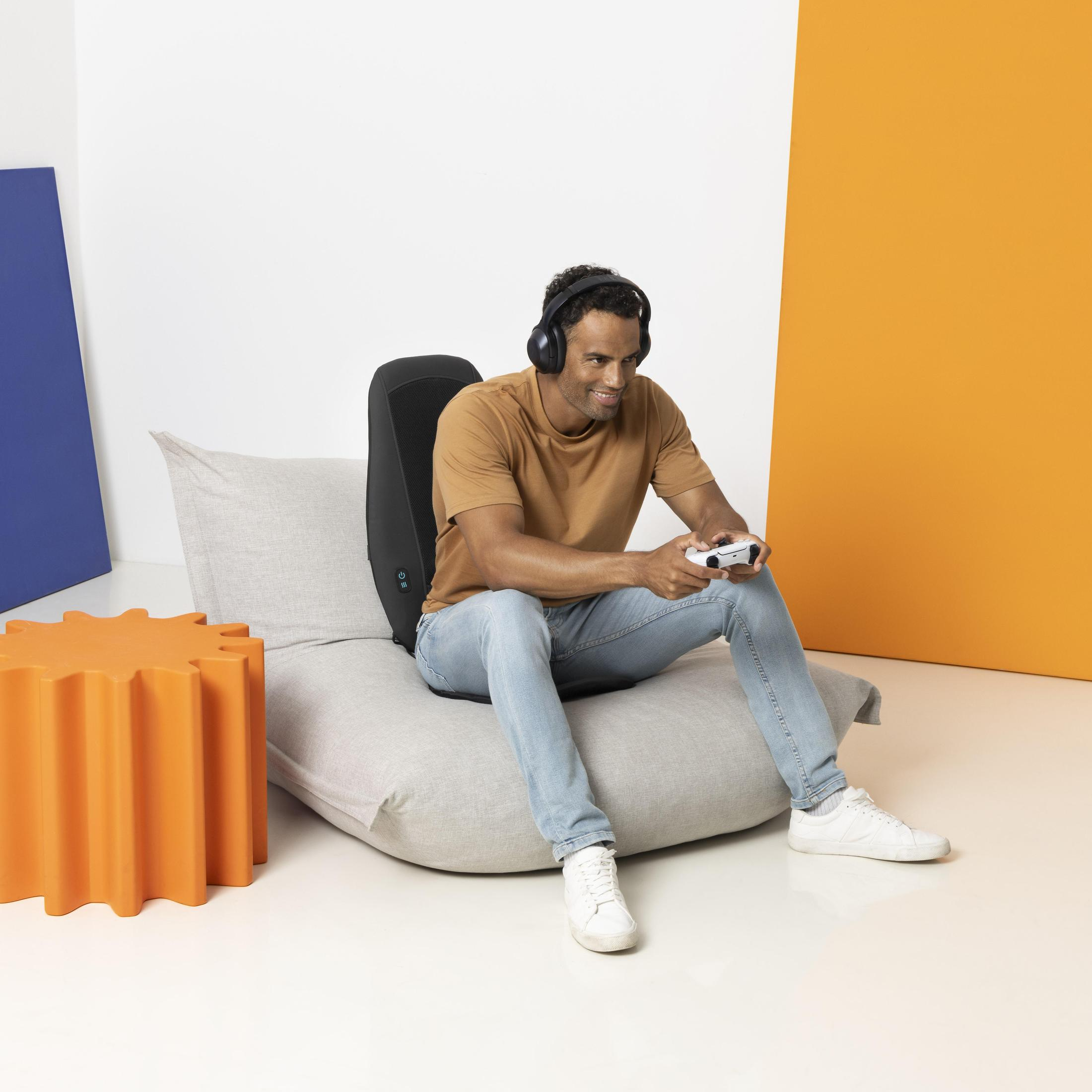 Mężczyzna siedzi, gra w grę za pomocą kontrolera, ma na sobie słuchawki i używa poduszki do masażu.