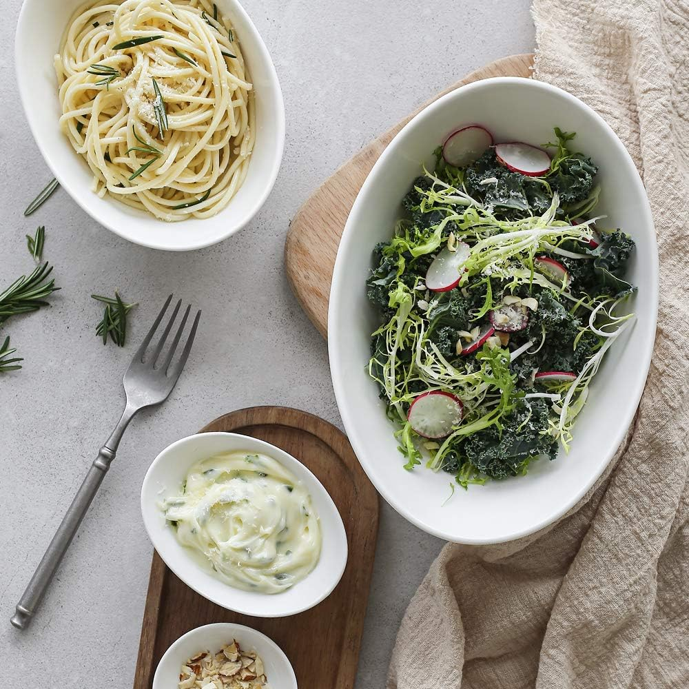Tre ciotole ovali bianche con cibo, tra cui pasta e insalata. Forchetta e vassoio in legno.