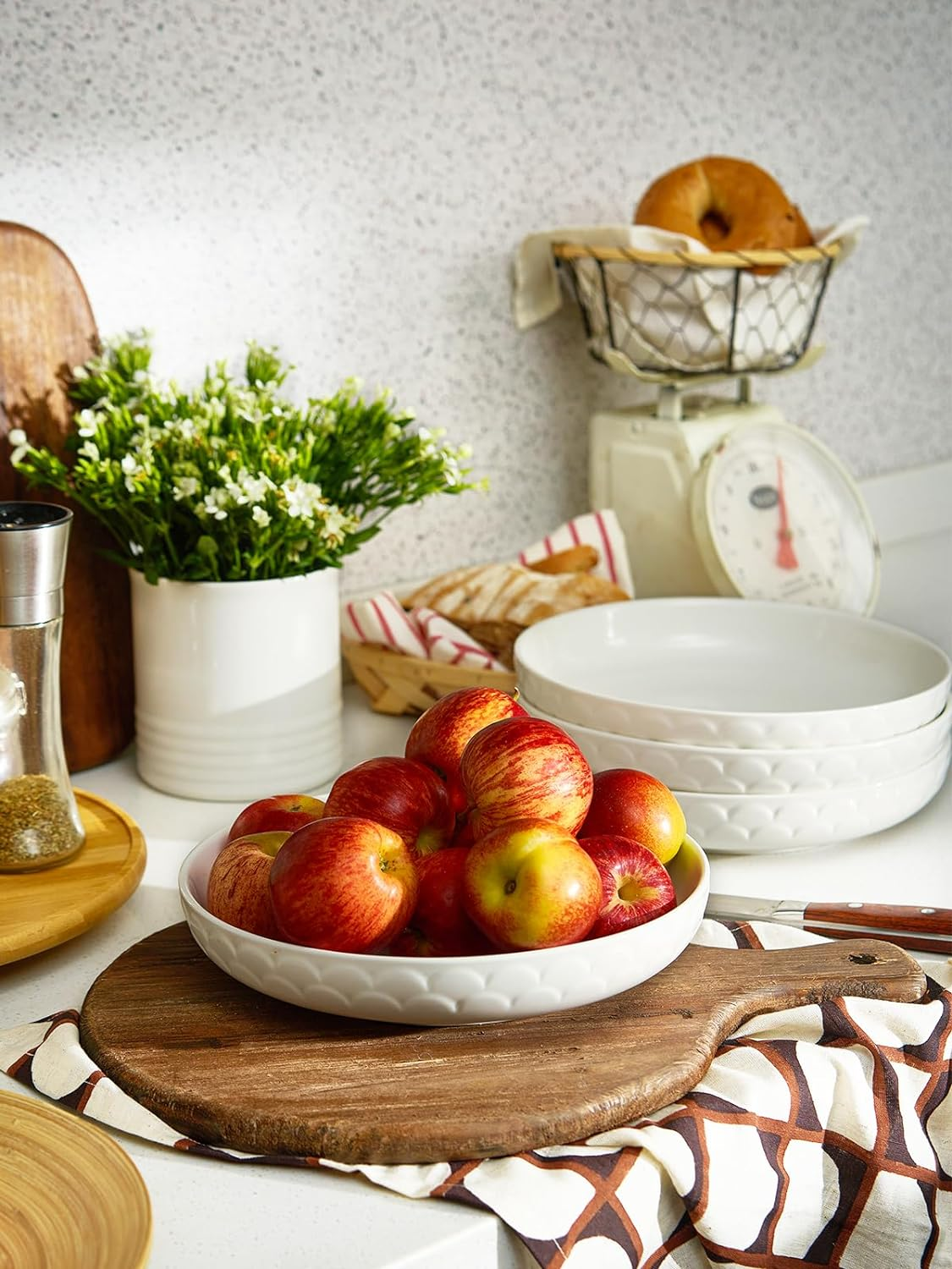 Una ciotola di mele rosse su un tagliere di legno con un vaso bianco di fiori e altri utensili da cucina.