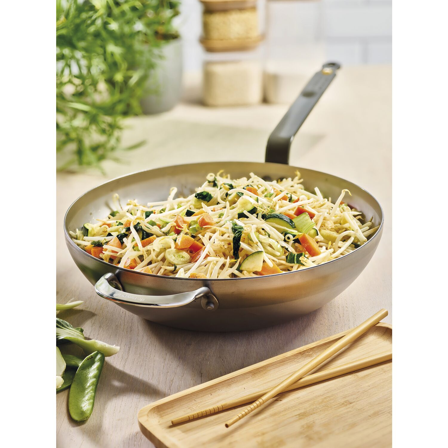 Ein Wok mit gebratenem Gemüse und Nudeln, Essstäbchen und ein Holztablett auf einem Tisch.