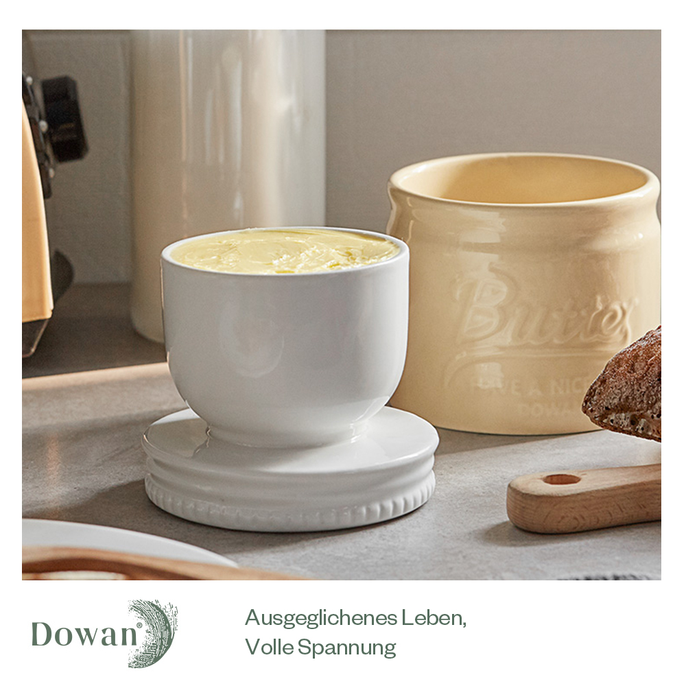 Ein weißes Buttergefäß und ein cremefarbenes Glas mit Butter auf einer Oberfläche.