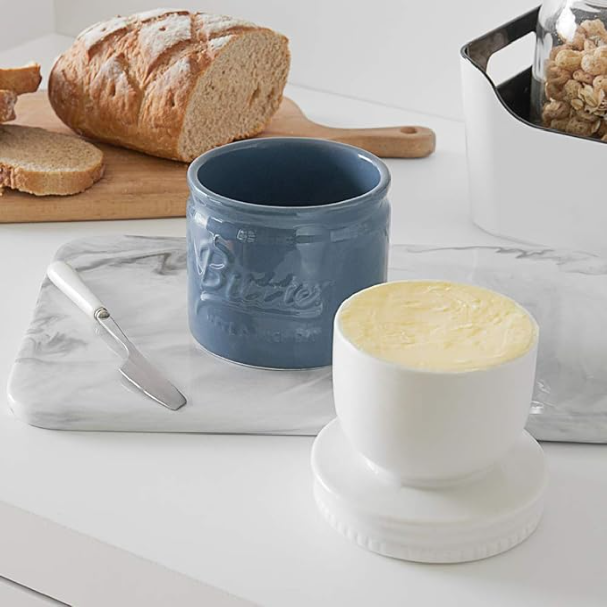 Butter mit Messer, blaue Butterdose, weiße Butterdose. Brot im Hintergrund.