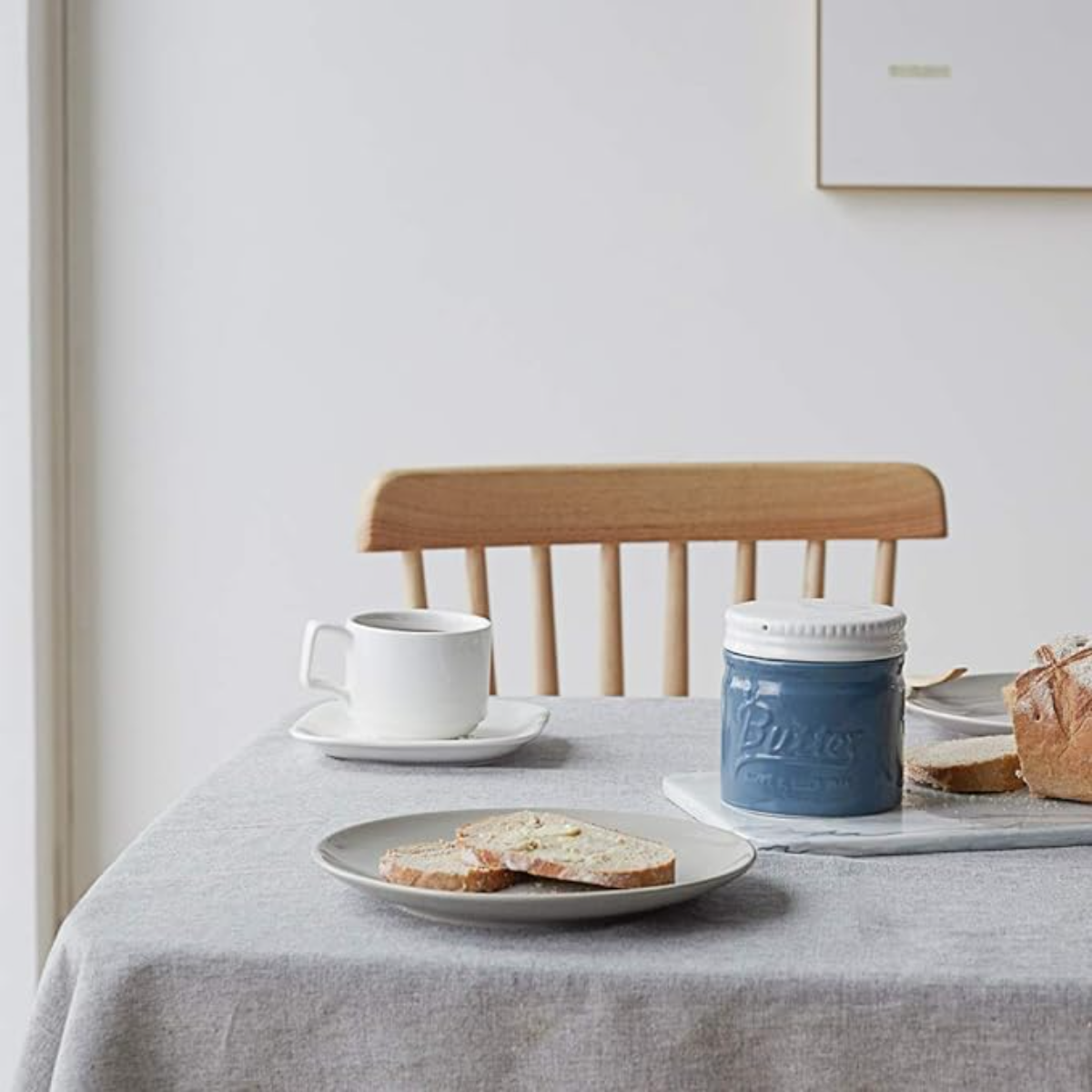 Ein Tisch mit einem Teller Brot mit Butter, einer Tasse und einer Butterdose. Ein Stuhl ist im Hintergrund.