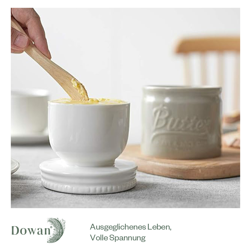Weiße Butterdose mit Butter und Holzmesser. Eine zweite Butterdose im Hintergrund.