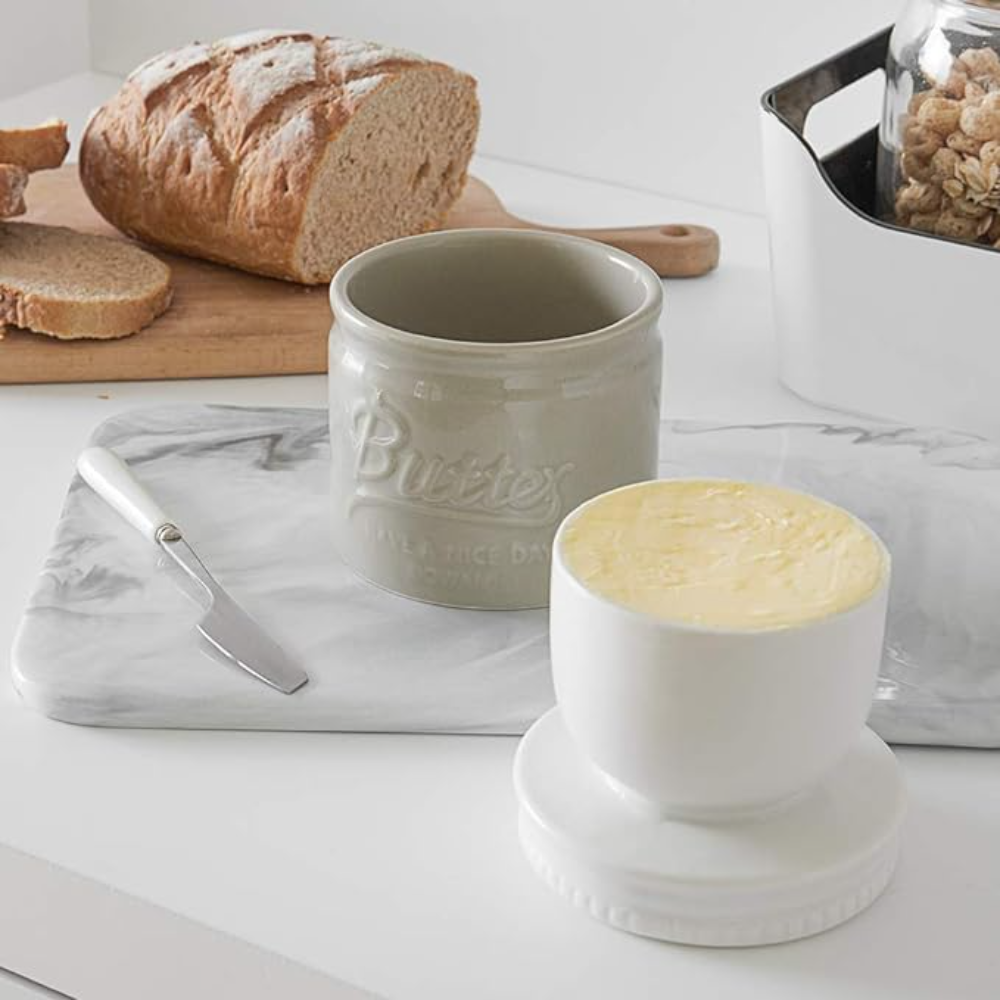 Ein graues Butterglas, eine Butterdose und Brot auf einer Marmoroberfläche mit Butter und Messer.