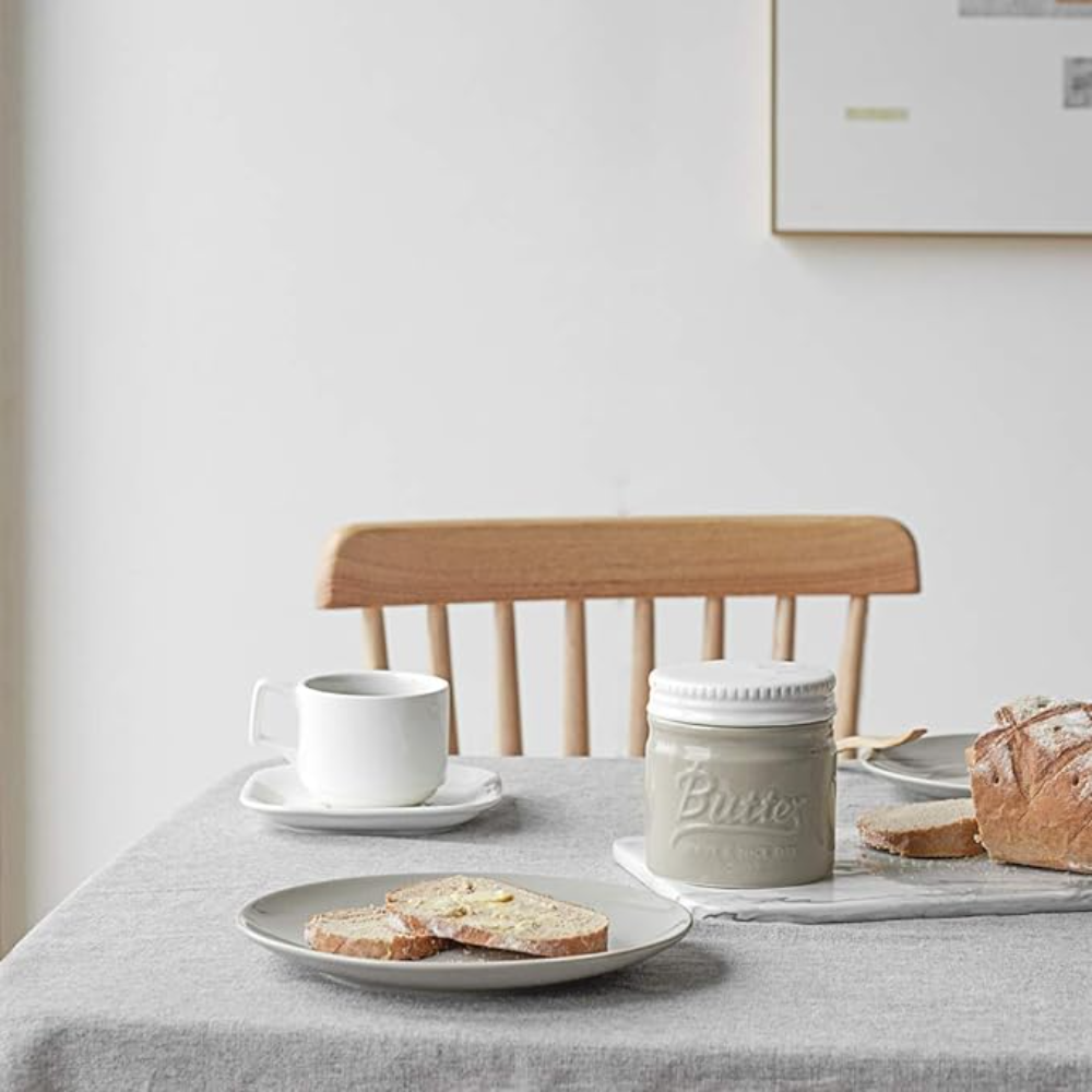 Tisch mit grauem Tischtuch. Enthält Teller mit Toast, Tasse und Butterdose.