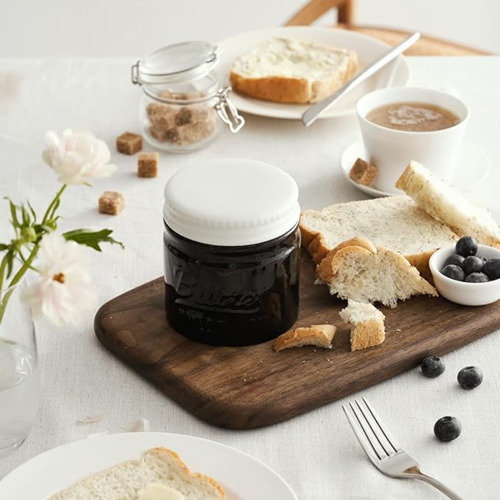 Un tavolo con burro, toast, un barattolo di marmellata, una tazza di caffè e fiori. La scena è luminosa.