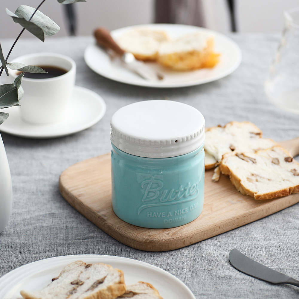 Ein türkisfarbenes Butterglas steht auf einem Holzbrett, umgeben von Brot und einer Kaffeetasse.