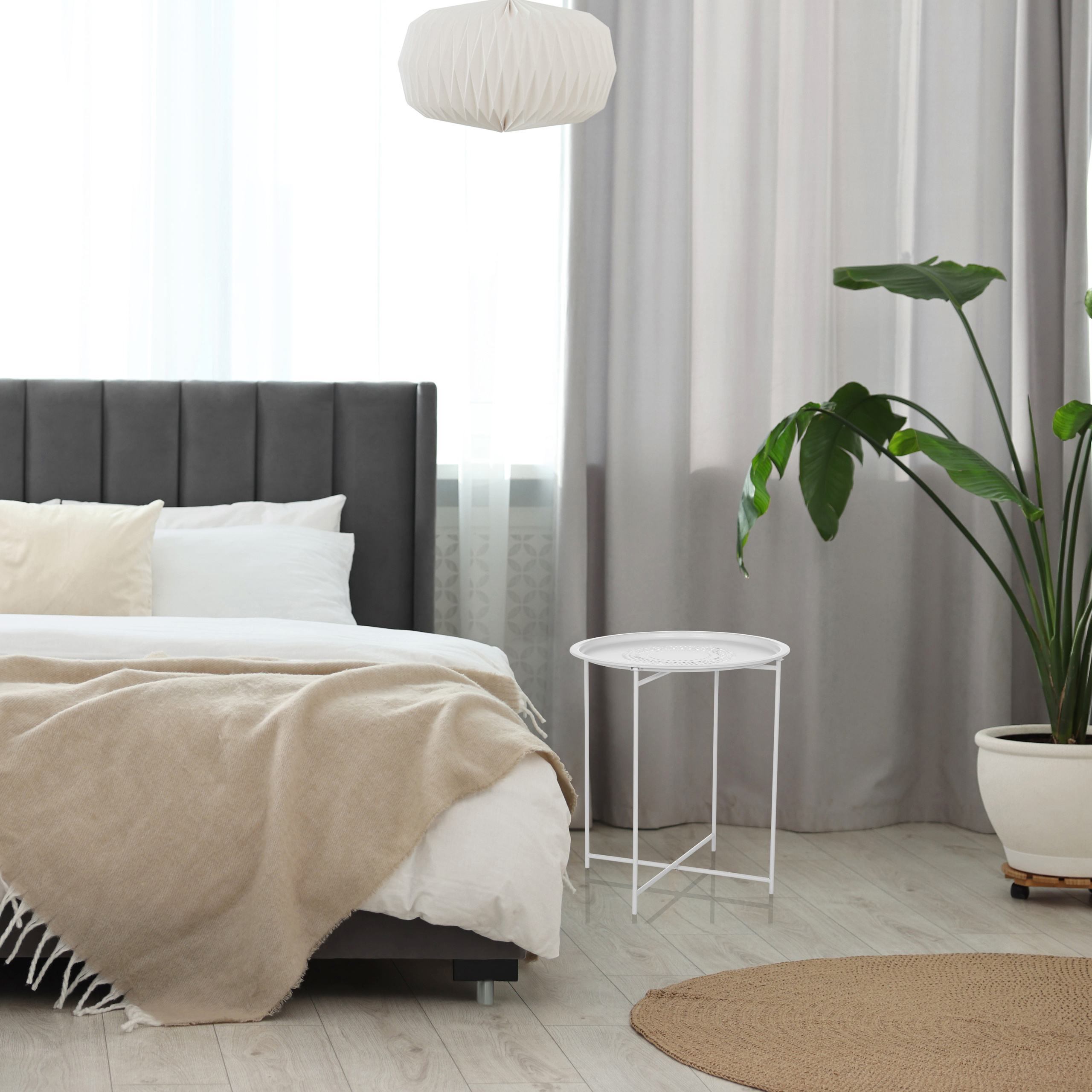 Ein Schlafzimmer mit Bett, Beistelltisch, Lampe, Vorhängen und einer Topfpflanze. Neutrale Töne schaffen eine ruhige Atmosphäre.