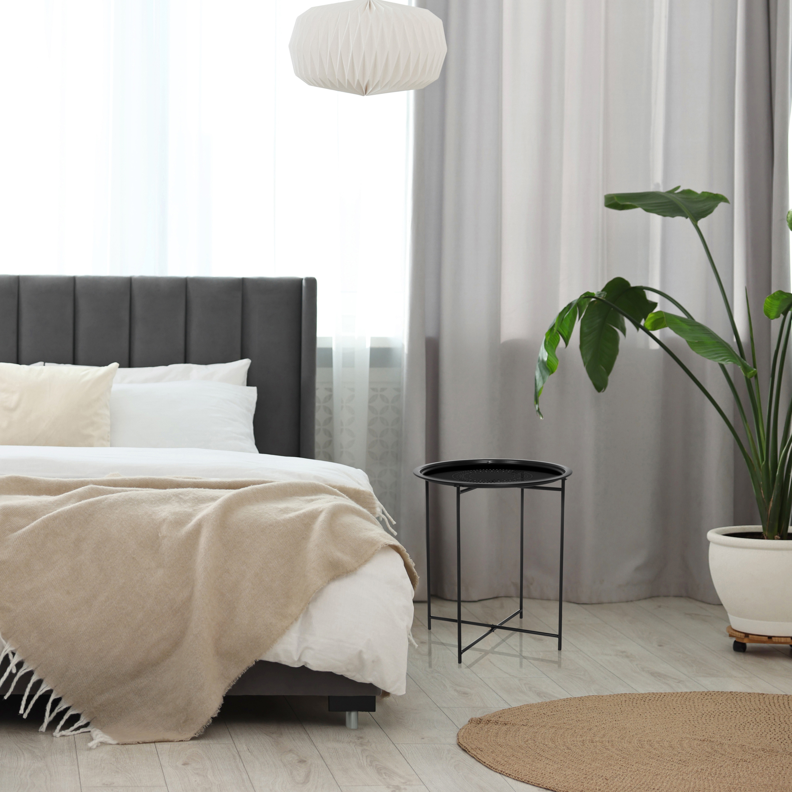 Schlafzimmer mit Bett, Pflanze, Beistelltisch und Vorhängen. Das Bett hat eine beige Decke. Ein weißer Lampenschirm hängt von der Decke.