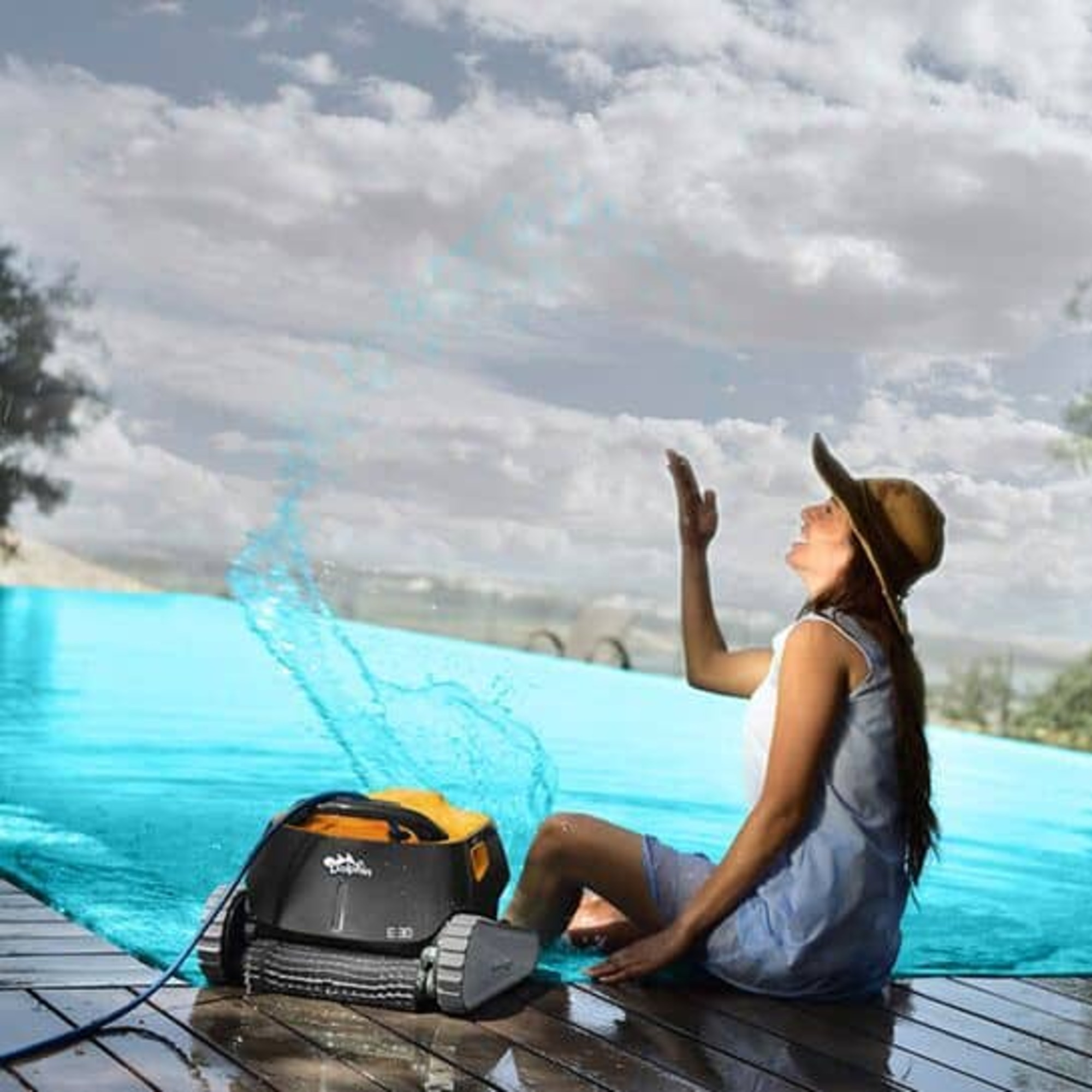 Mujer sentada junto a un limpiador de piscina, salpicaduras de agua. Cielo con nubes.
