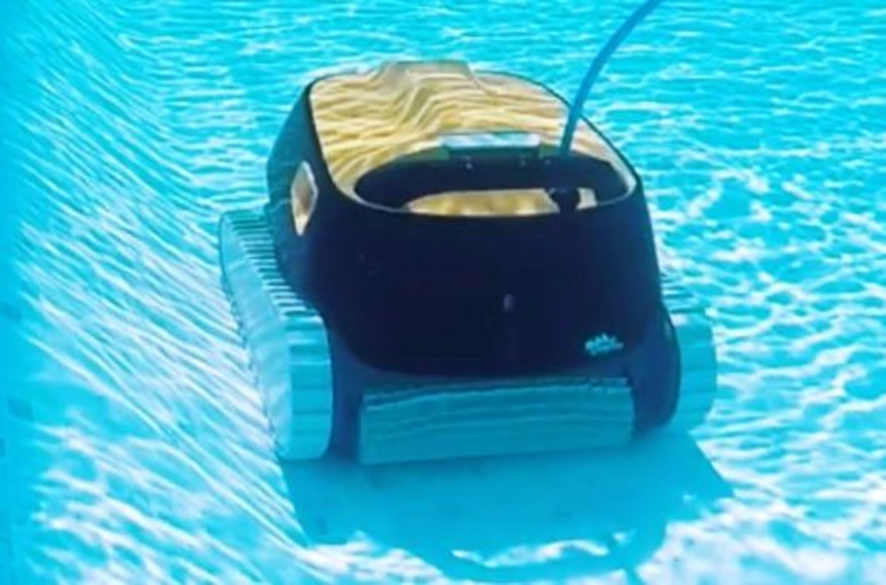 Un limpiador de piscinas en una piscina, con orugas y una parte superior oscura. El agua es clara con ondulaciones.